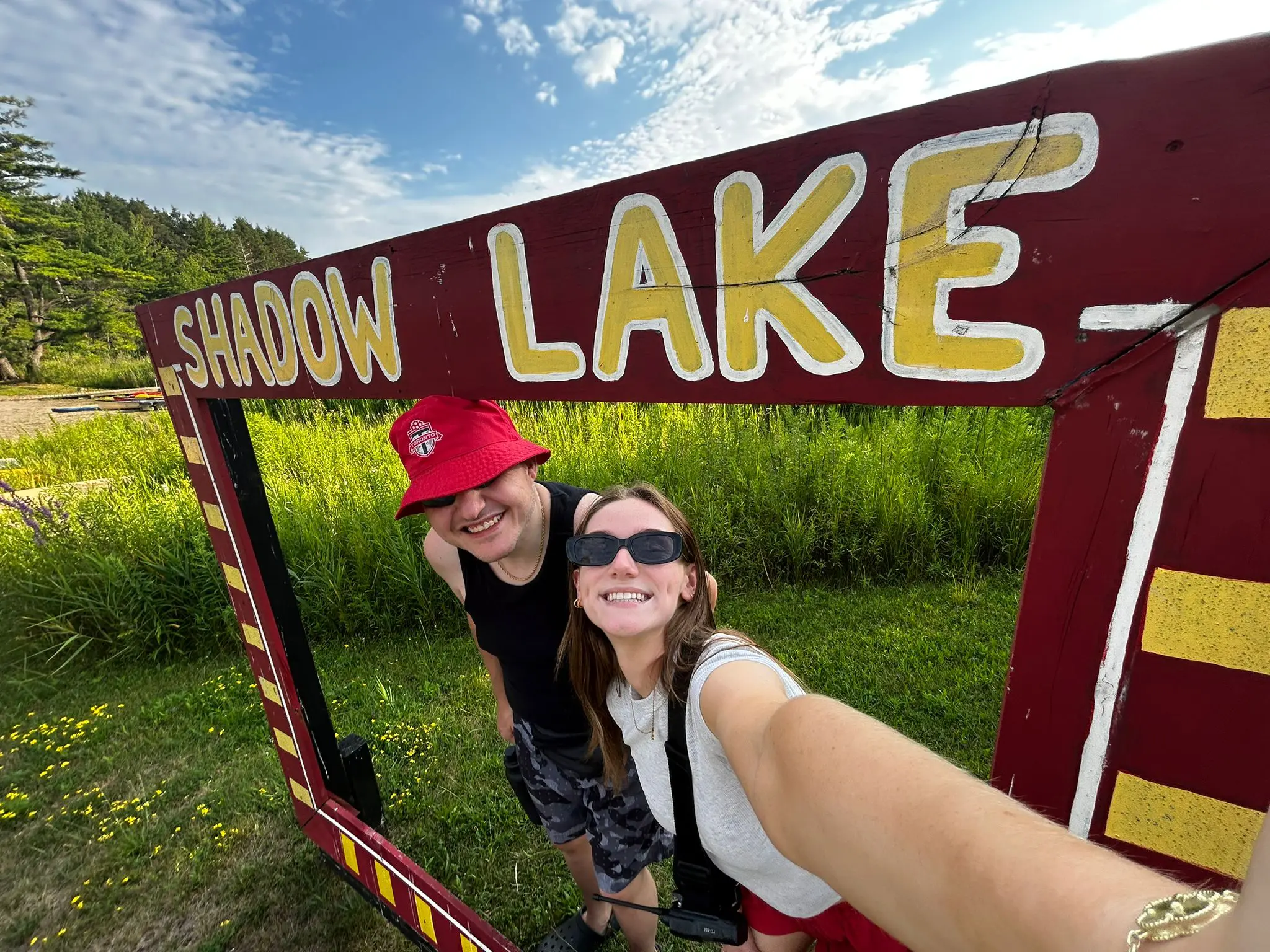 Two people smiling outdoors while posing inside a large Shadow Lake sign frame.