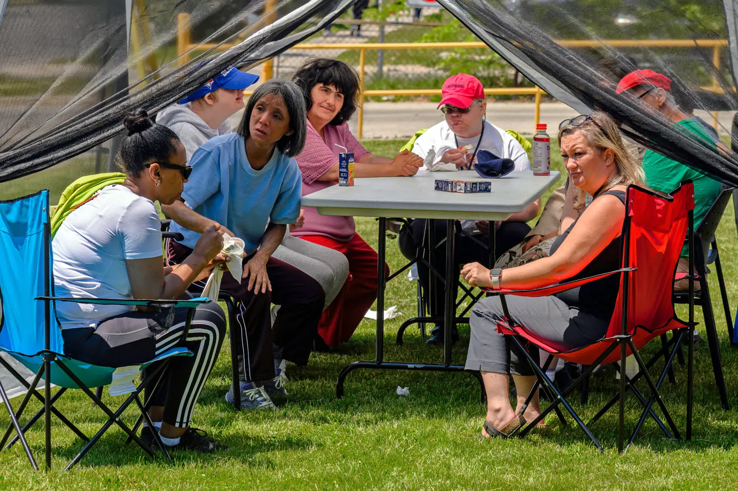 Community members sit under a shaded tent around a table during an outdoor event, socializing and taking part in activities on a sunny day.