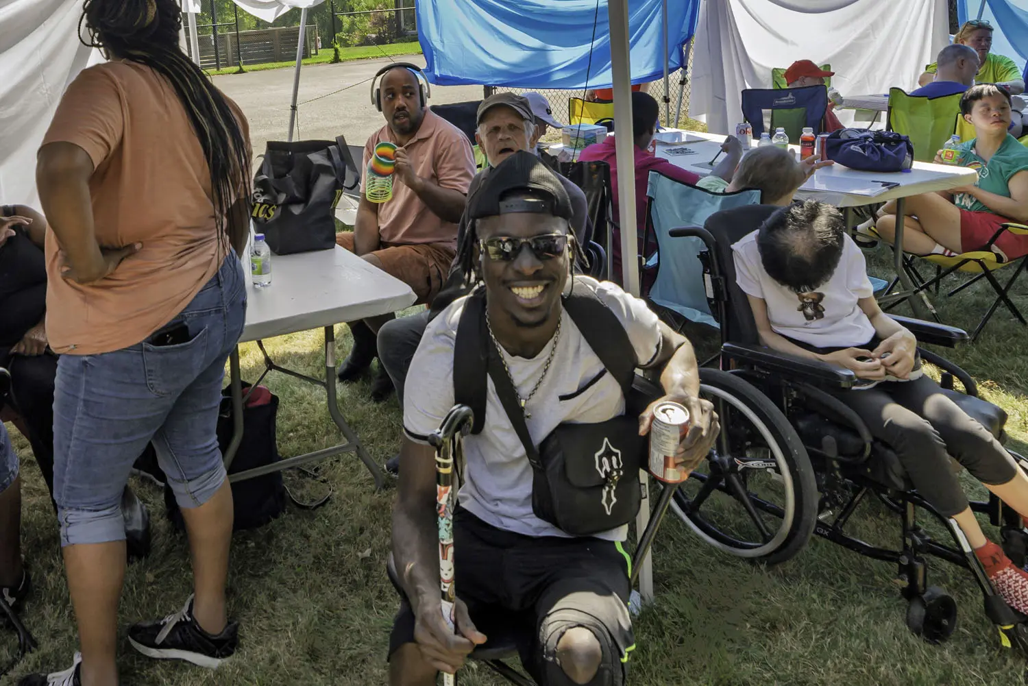 Community members gather under a tent at an outdoor summer event, participating in activities, relaxing, and connecting with others at tables and seating areas.