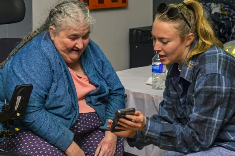 A person seated in a wheelchair and another person sitting beside them look at a smartphone together in an indoor community setting.