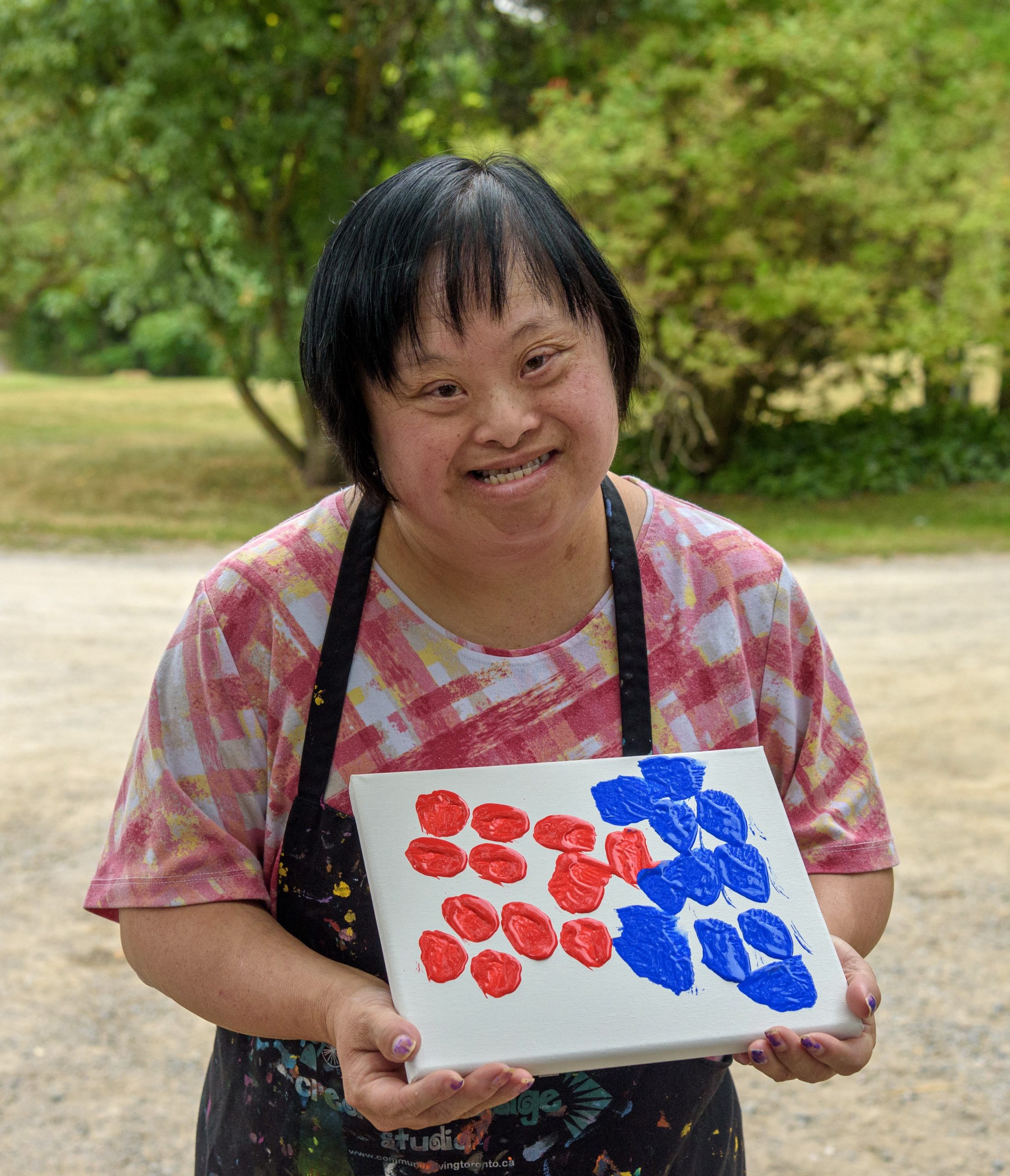 A woman with short black hair smiling while proudly holding up her artwork she created. Artwork is all white canvas with 12 red dots on one side and 11 blue dots on the other side. 