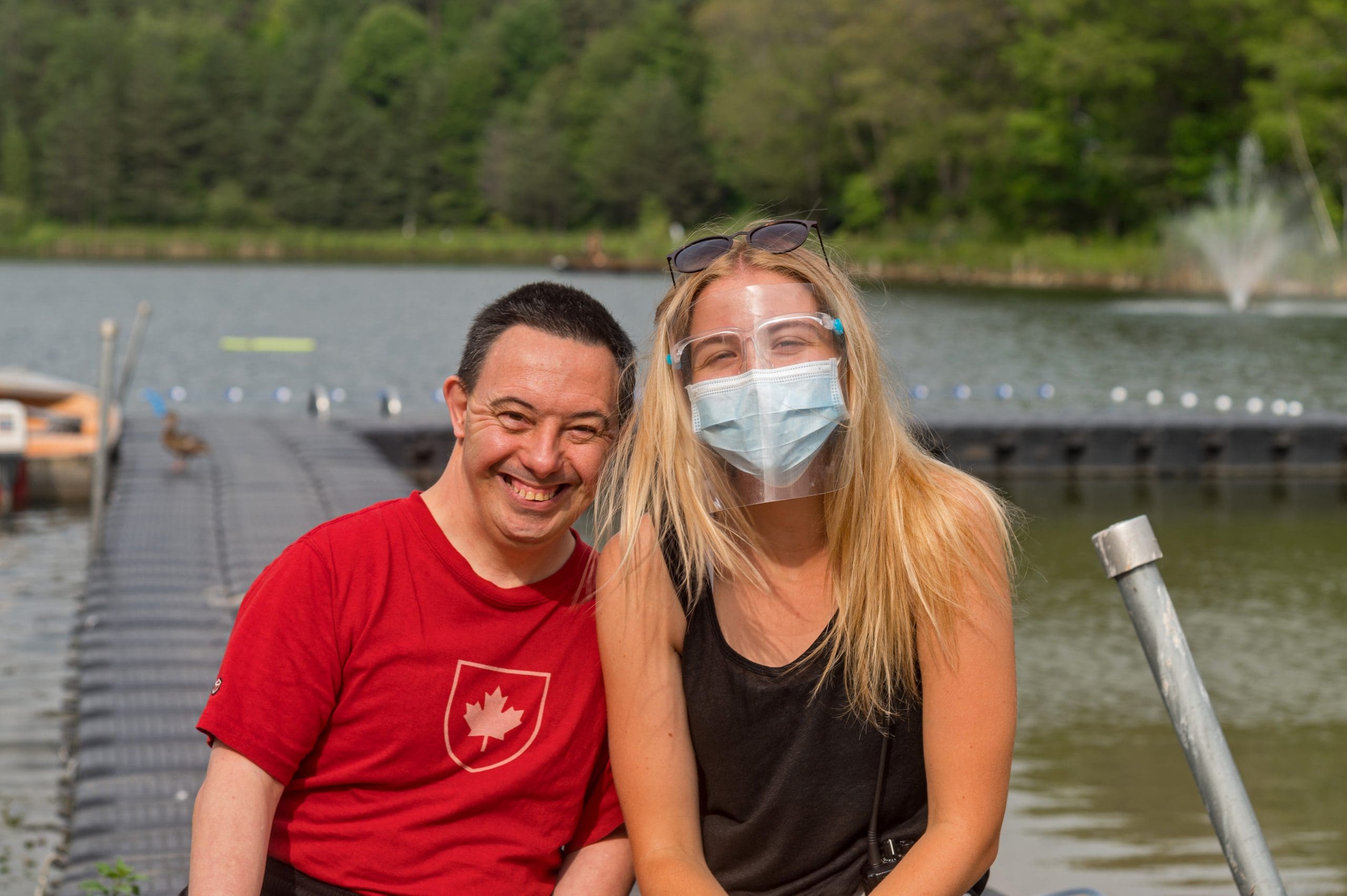 A camper and staff sitting in front of Shadow Lake in the sun. Staff member wearing a mask