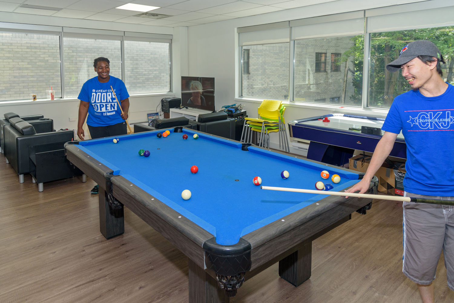2 people wearing 2 different blue T-shirts playing pool. The pool table is also a similar blue as the T-shirts