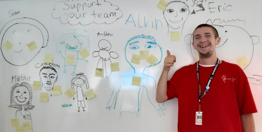 A guy with a red T-shirt standing infront of a whiteboard. Many fun stick figure drawings on the whiteboard.