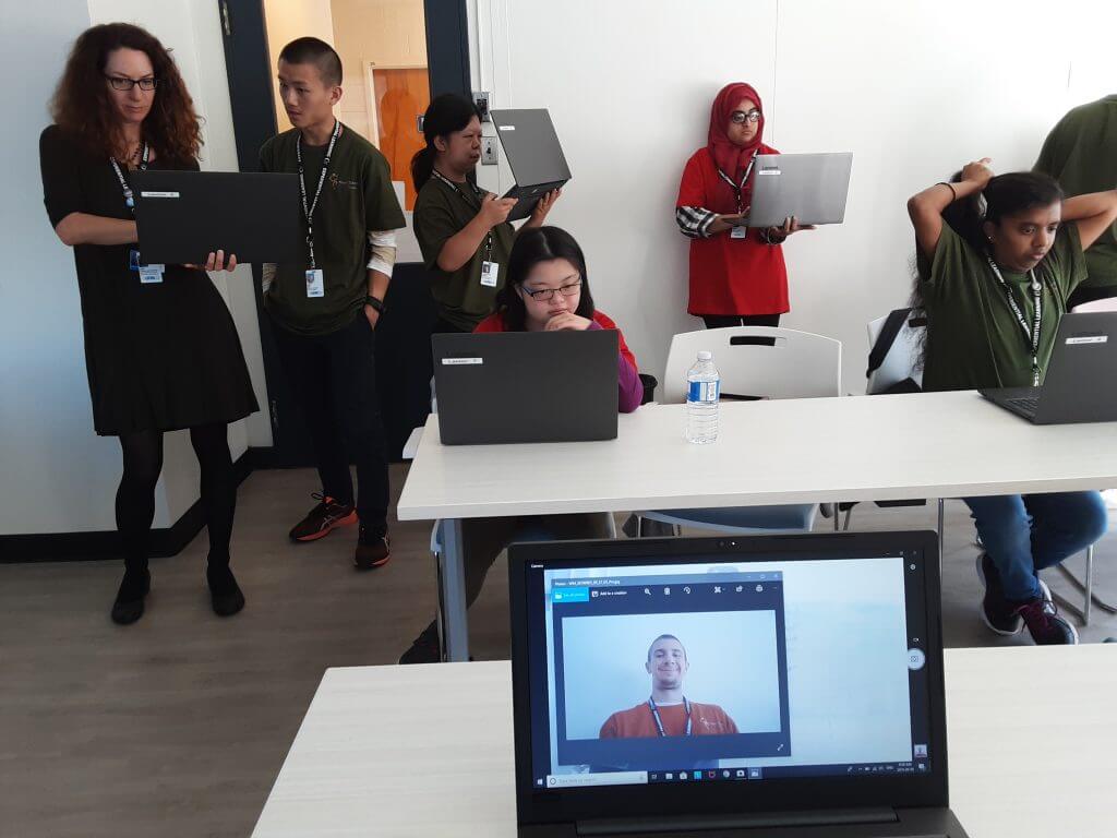 Students and instructor in a class room. MOst people standing holding their own laptop, 2 people sitting at the 2 desk/tables with their laptops