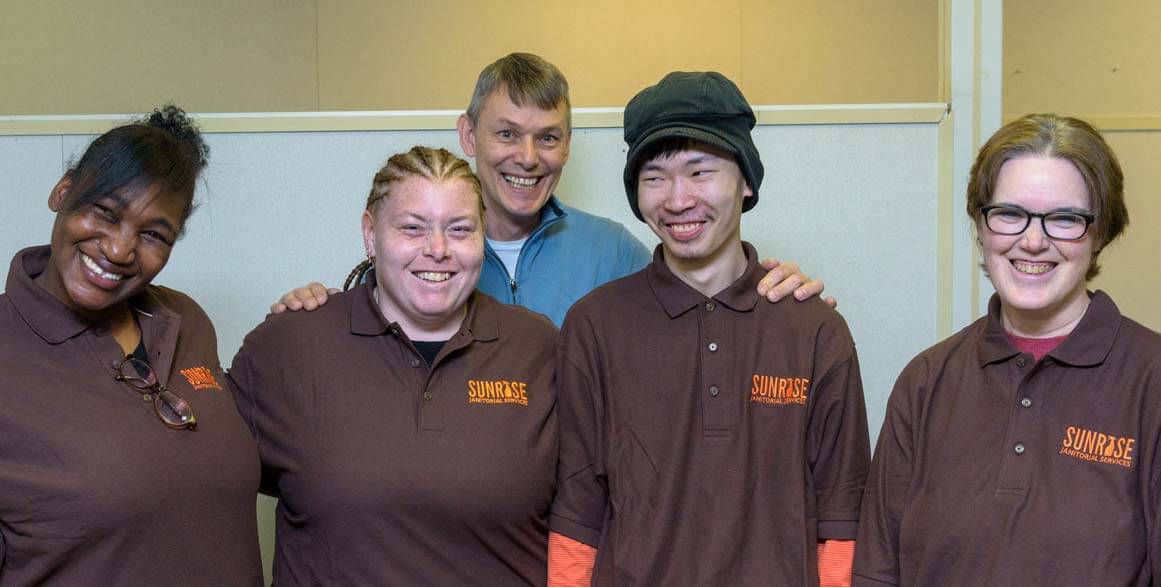 Sunrise Janitorial Staff photo. 4 people standing together with brown sunrise Shirt uniform and Rodney behind the group of 4 wearing a blue shirt.