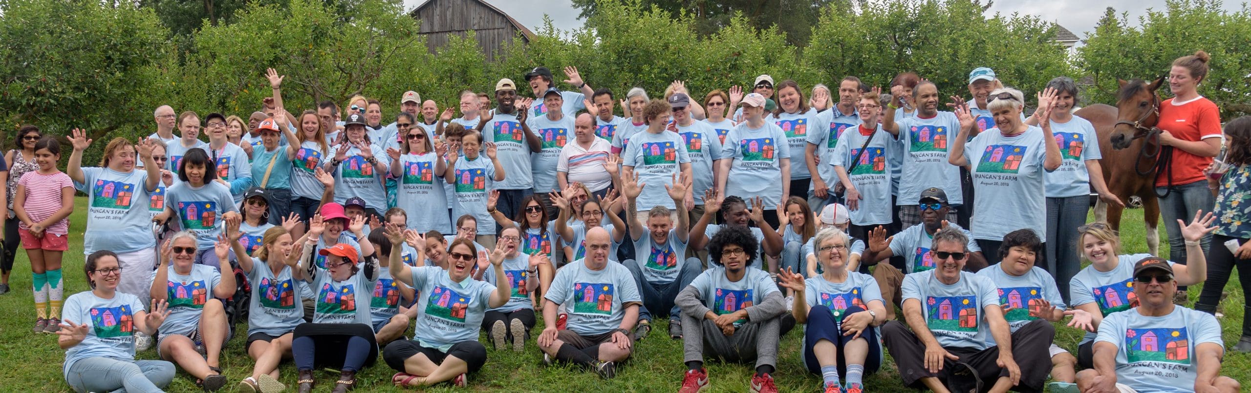 A huge group of people sitting and standing together wearing the same blue T-shirt with a colourful painting of a farm scene. Everyone doing jazz hands. A brown horse is also in the photo on the right of the group