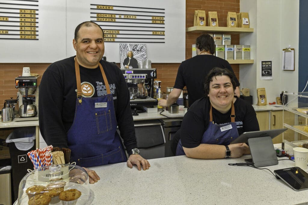 Here are 2 baristas behind the counter at the social brew. Smiling one is standing and one is leaning on the counter, casual. There is a person behind them with their back to the camera