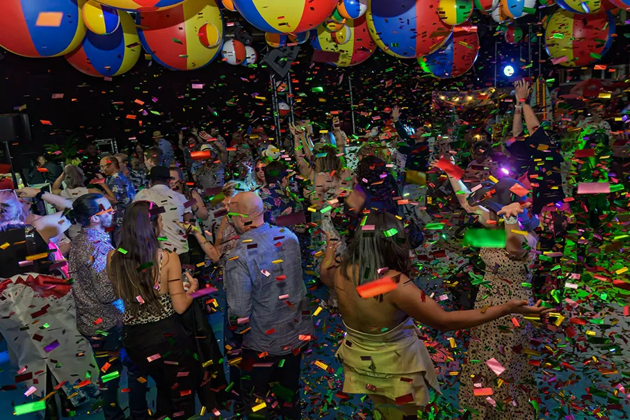 A crowd celebrates at a lively indoor party as colourful confetti falls beneath giant beach balls and bright stage lighting.
