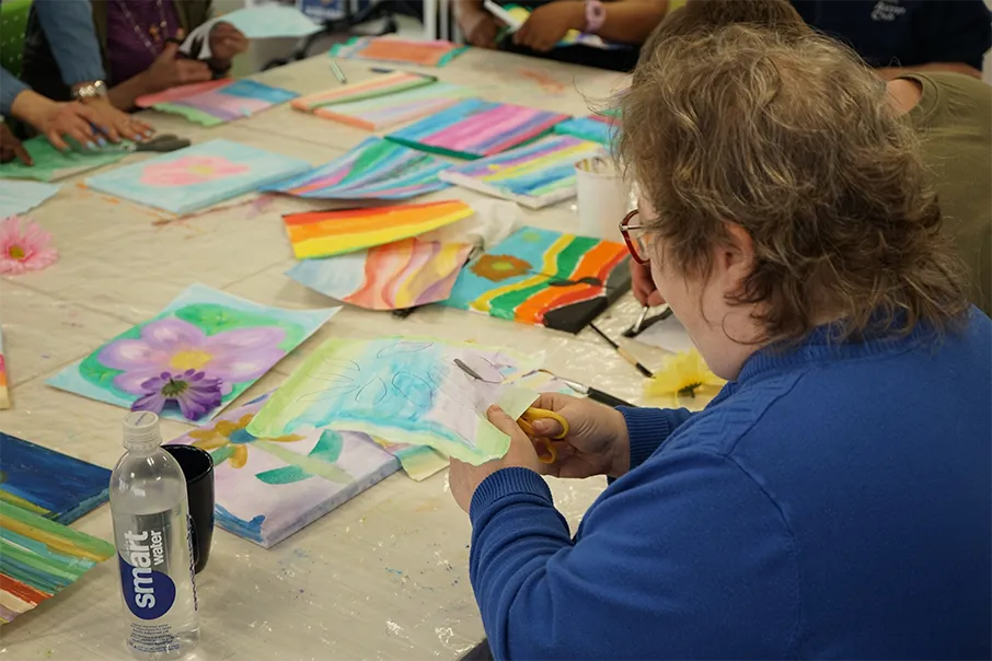 A person in a blue sweater cuts paper at an art workshop with a table covered in colourful paintings and craft supplies.