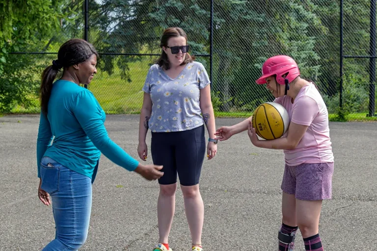 Three people standing on an outdoor court, with one person in a pink helmet holding a basketball during a group activity.