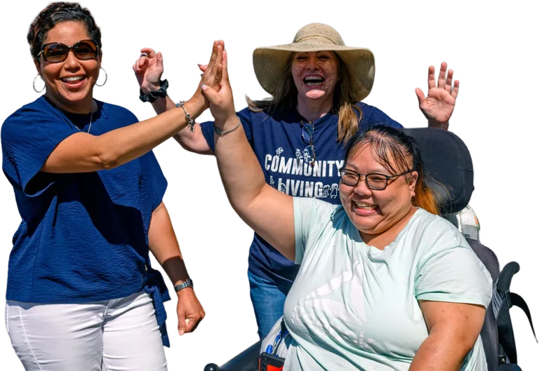 Three people smiling and high-fiving outdoors at a Community Living event, including tenants and community members in the background.