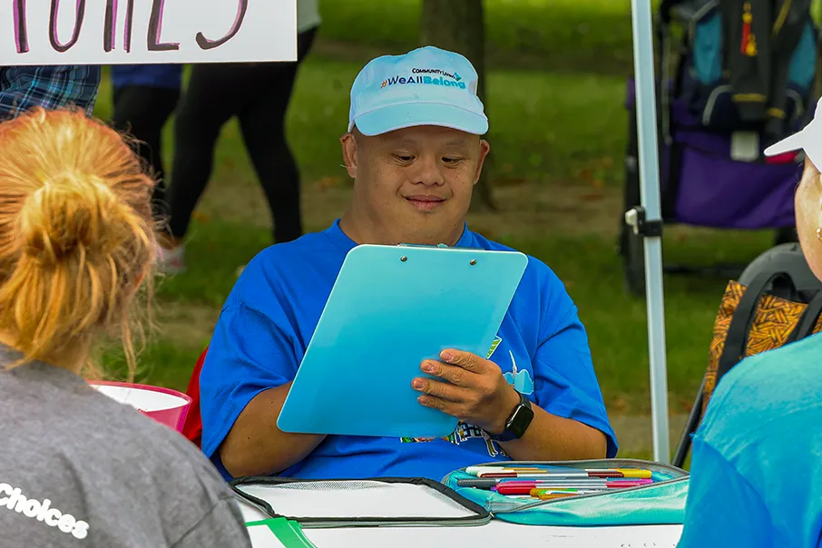 Person seated at a community event, writing on a clipboard while participating in an activity and engaging with others nearby.