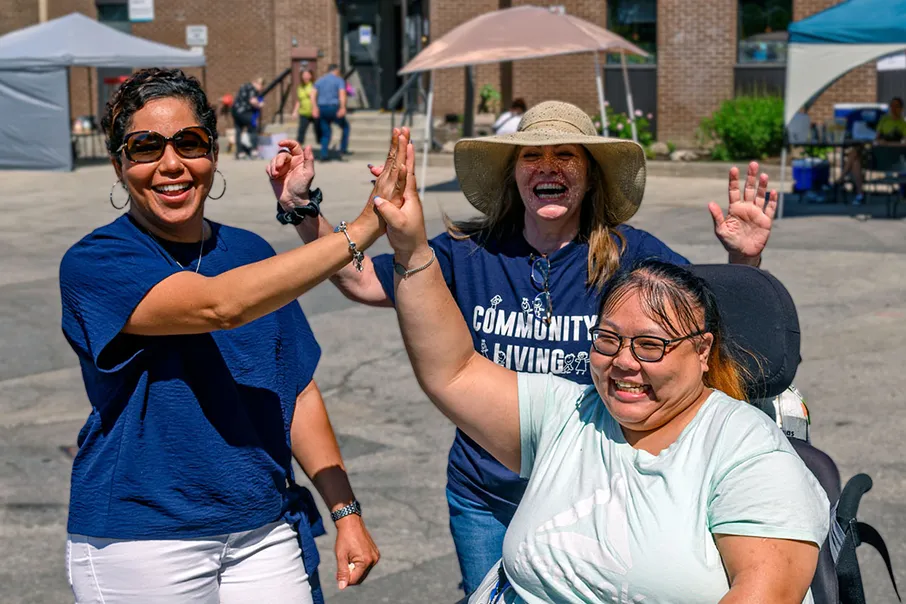 Three people smiling and high-fiving at an outdoor Community Living event, celebrating connection, inclusion, and community spirit.