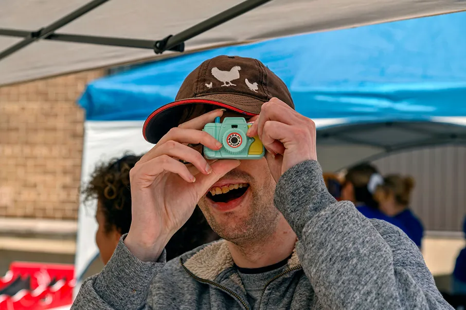 Person smiling while taking a photo with a toy camera at an outdoor community event under a tent.