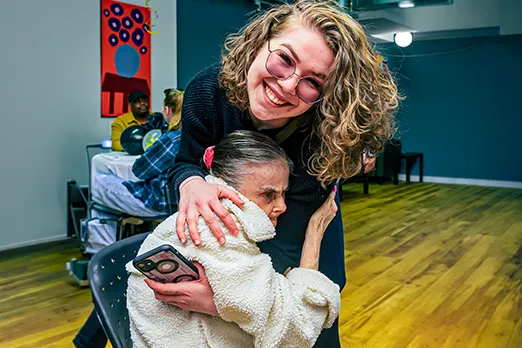 A woman with curly hair and glasses warmly hugging a woman in a cozy indoor community space.