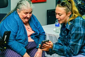 A woman with glasses shows something on her phone to the woman in a wheelchair.