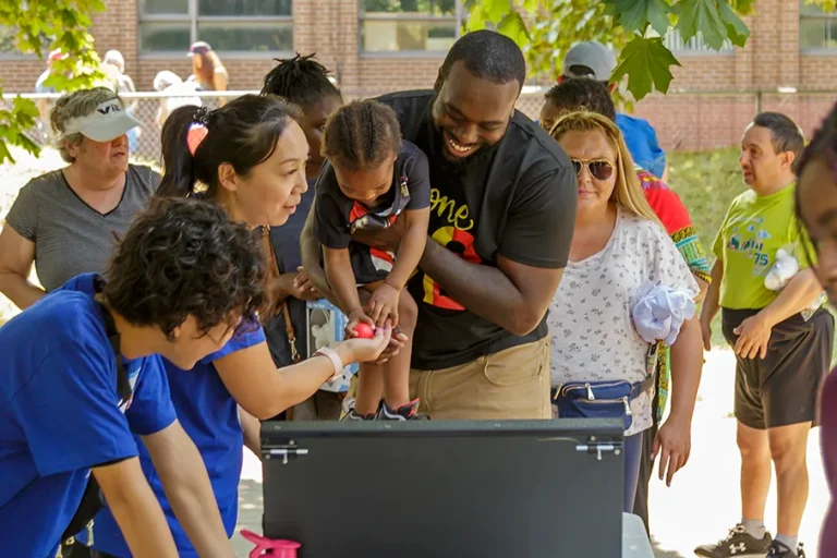 Families and community members gathered outdoors, helping a child with an activity at a Community Living event.