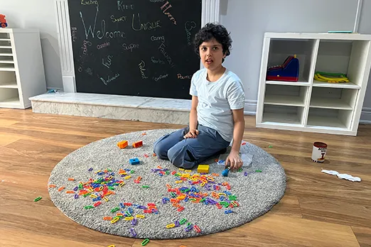 A child sitting on a round rug surrounded by colourful paper clips in a bright classroom with a chalkboard wall behind them.