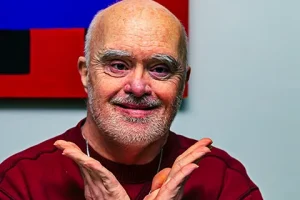 A smiling man in a red sweater poses indoors with hands open beneath his face.