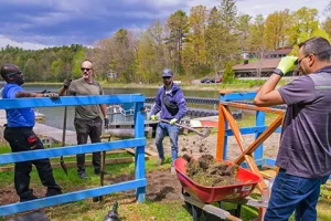 Four people work together outdoors near a lake, using tools and a wheelbarrow.
