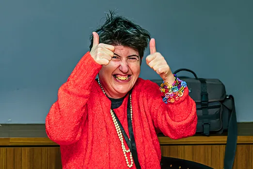 A person in a bright red sweater smiling and giving two thumbs up while wearing colourful bracelets and a pearl necklace.