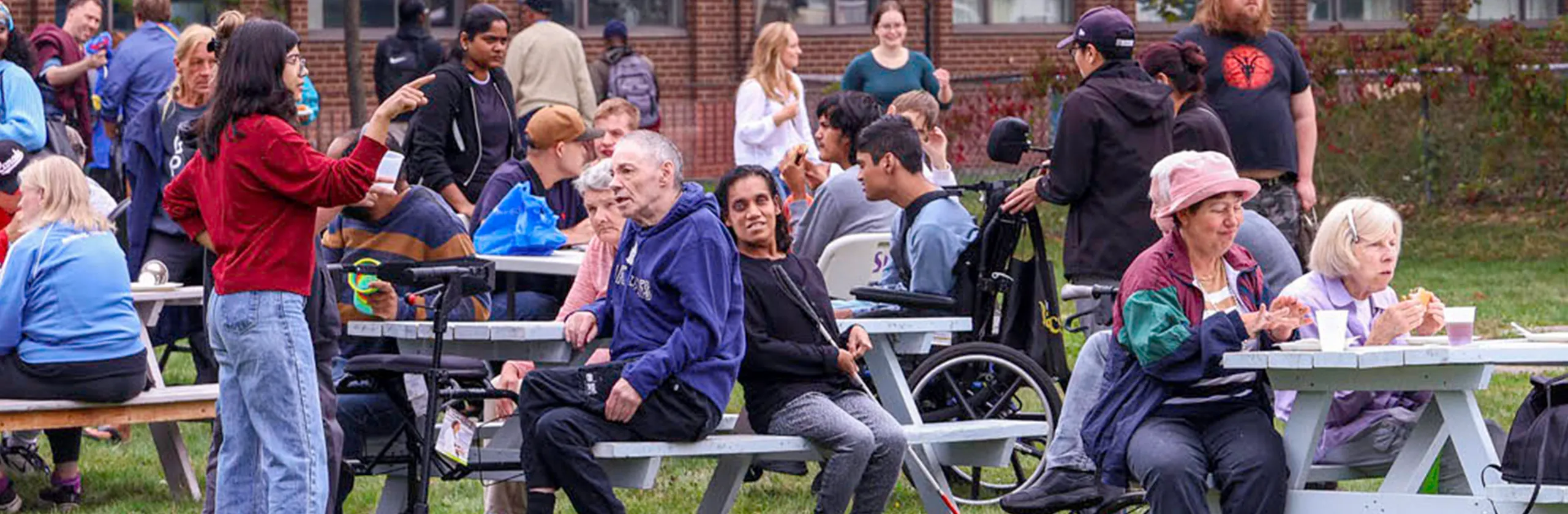 A large group of people gathered outdoors at picnic tables, socializing and enjoying a community event on a lawn.