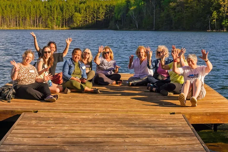 A group of people sitting on a dock by the lake, smiling and waving together during an outdoor gathering.