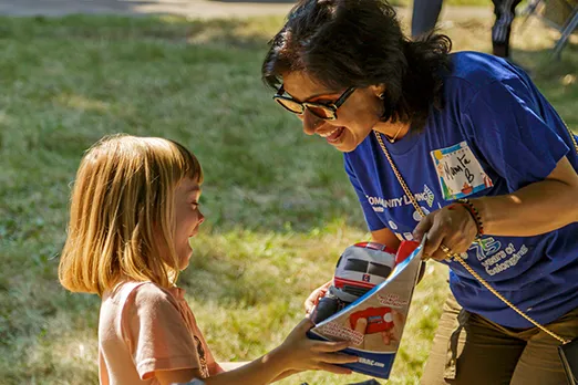 A woman in a blue shirt smiling while handing a toy train set to a young child.