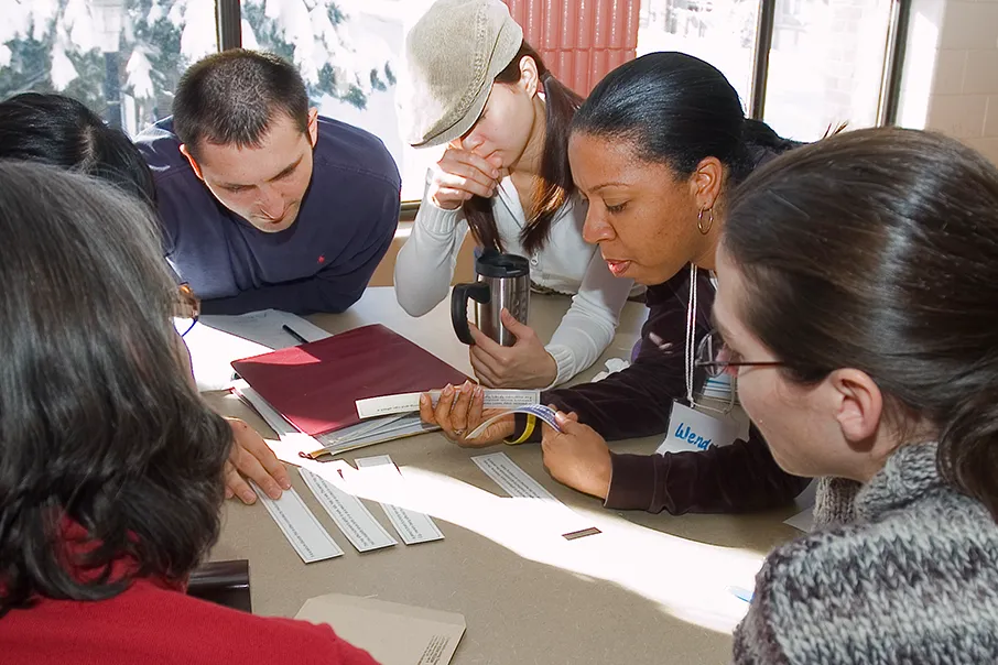 A group of people gathered around a table, reading and discussing printed materials during a collaborative workshop session.