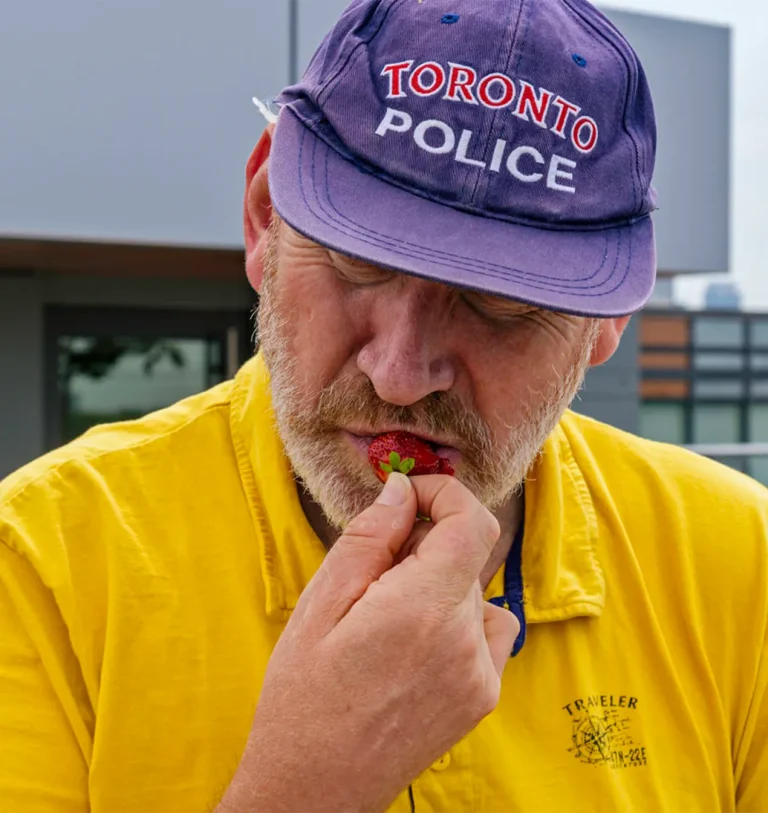 Close-up of a man in a Toronto Police cap and yellow shirt eating a fresh strawberry outdoors.