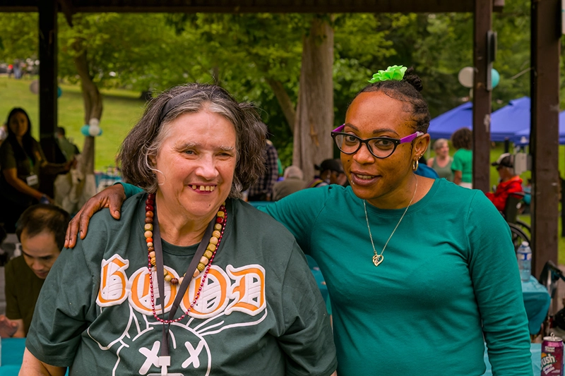Two community members stand together under a pavilion at an outdoor event, with tables and attendees in the background.