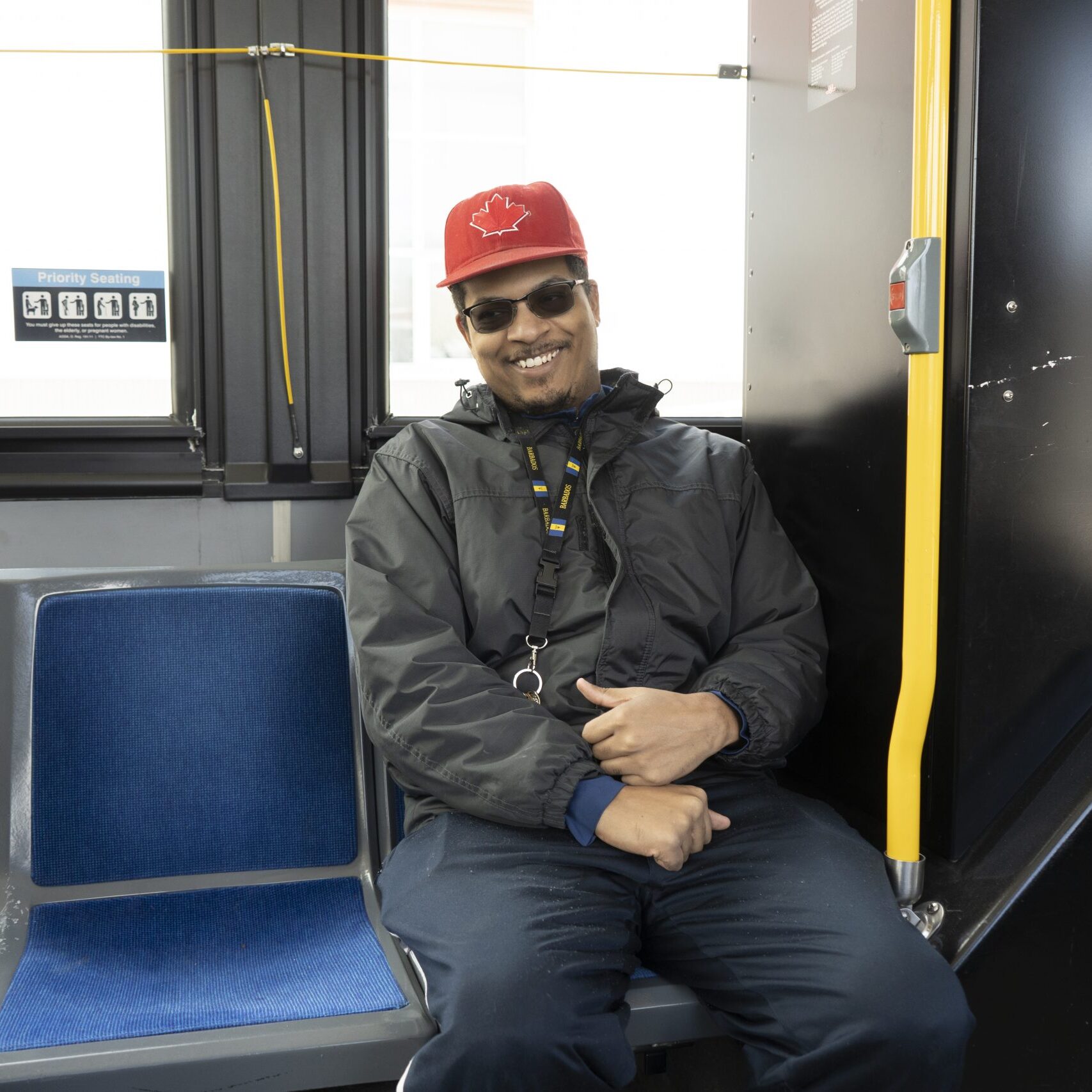 Josh smiling, sitting on a TTC bus seat facing inside the bus. Wearing sunglasses, a red maple leaf hat and a jacket.
